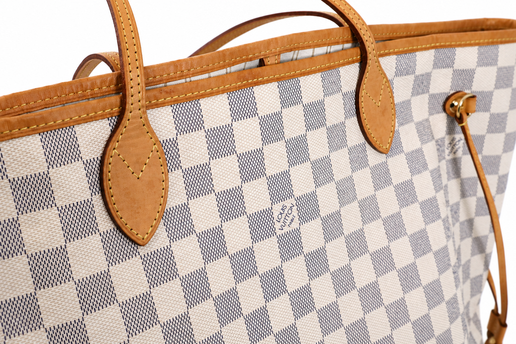 Close-up of a Louis Vuitton tote bag featuring the brand's signature Damier Azur canvas checkered pattern in beige and blue-gray, accented with tan leather trim and handles.