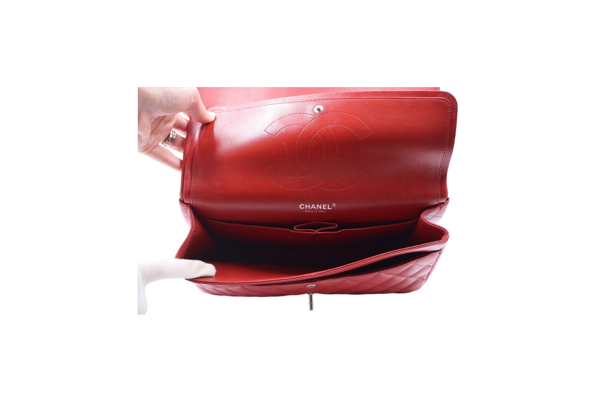 interior view red Chanel shoulder bag shown open, with a hand holding the top flap and a white-gloved hand pulling the bottom flap. The interior features a quilted leather construction, a CC logo embossed on the inside flap, and the Chanel name printed in white.