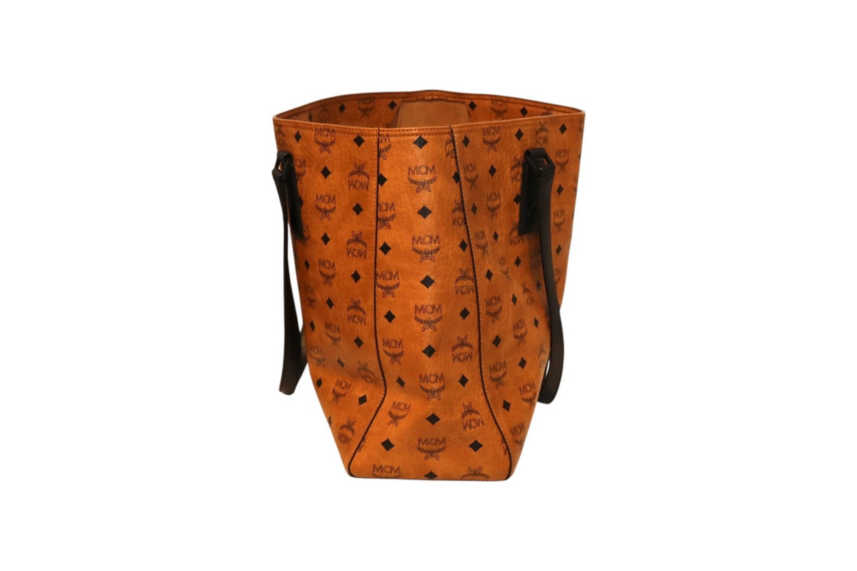 Side view of MCM orange Visetos tote bag with black monogram print, black leather shoulder straps, and an open-top design on a white background.