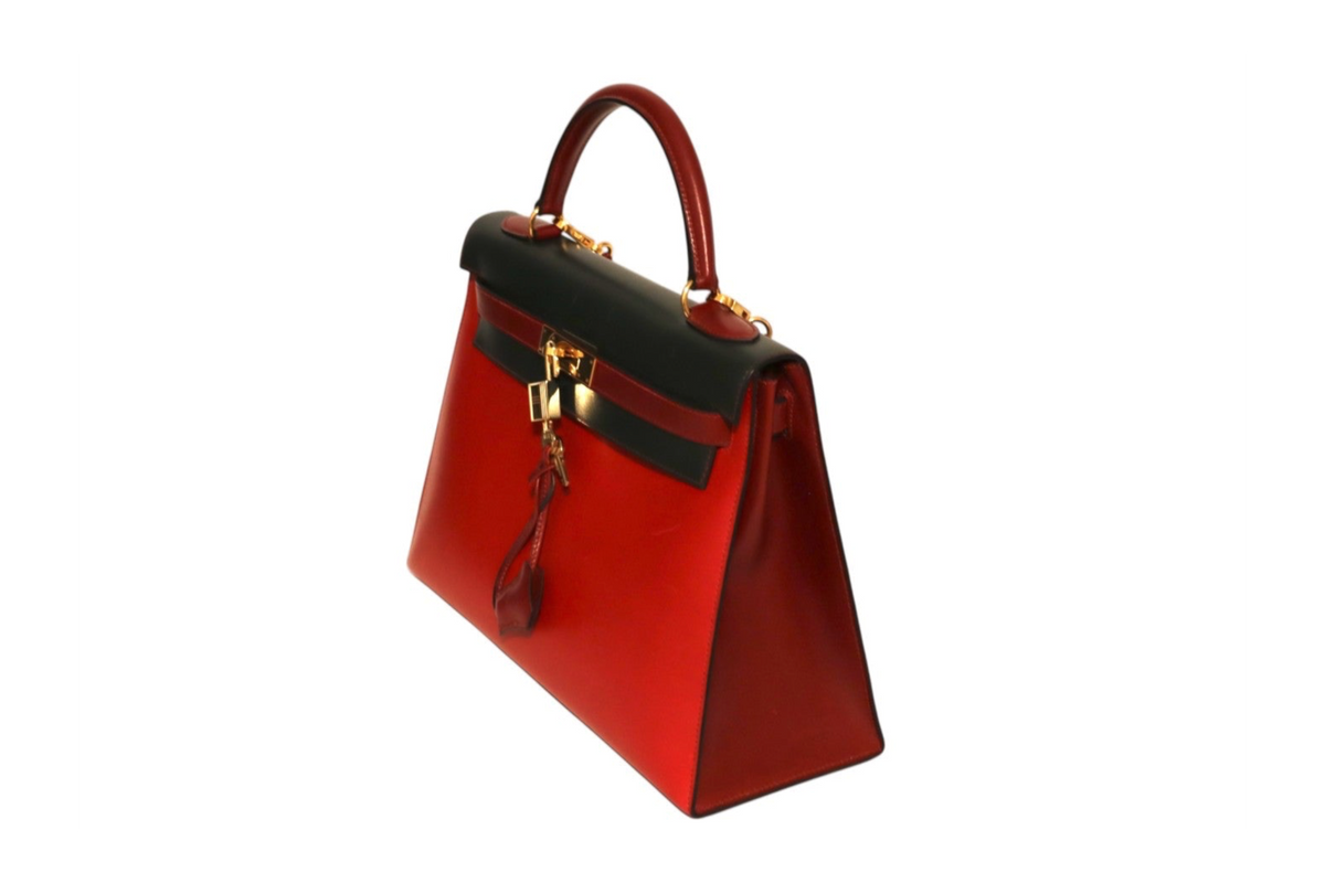 A structured Hermes red leather handbag with a black leather top handle and trim, gold-tone hardware, and a hanging clochette, shown from an angle.