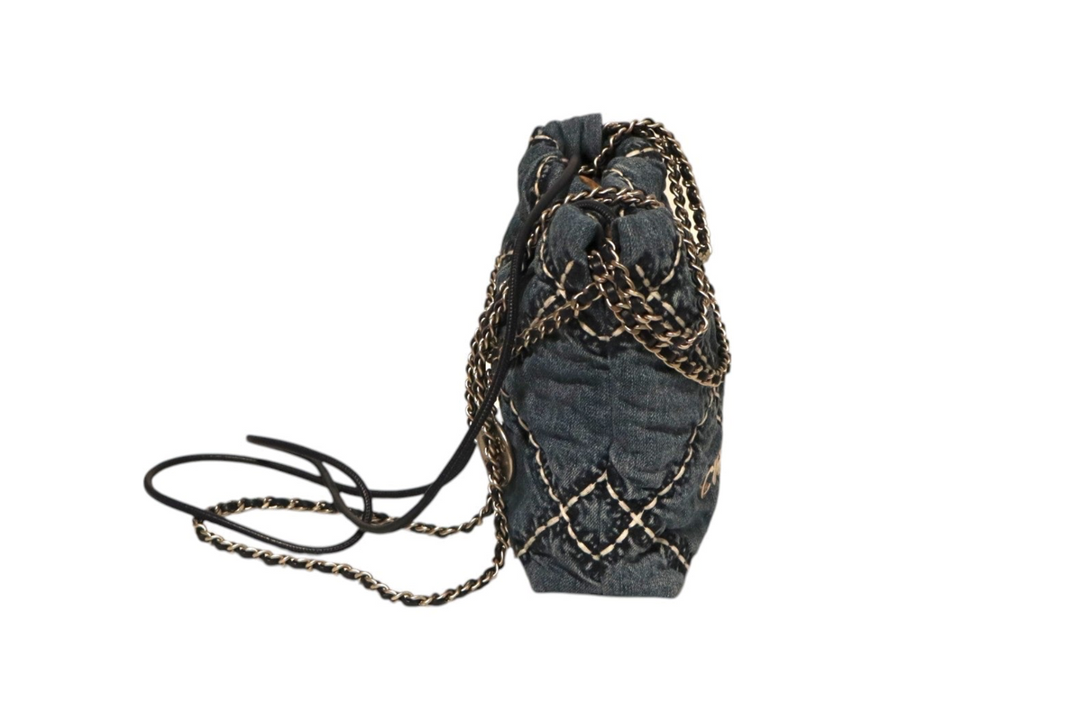 Side view of Chanel dark denim quilted drawstring bucket bag with gold-tone chain detailing, black leather drawstrings, and long leather-and-chain shoulder strap on a white background.