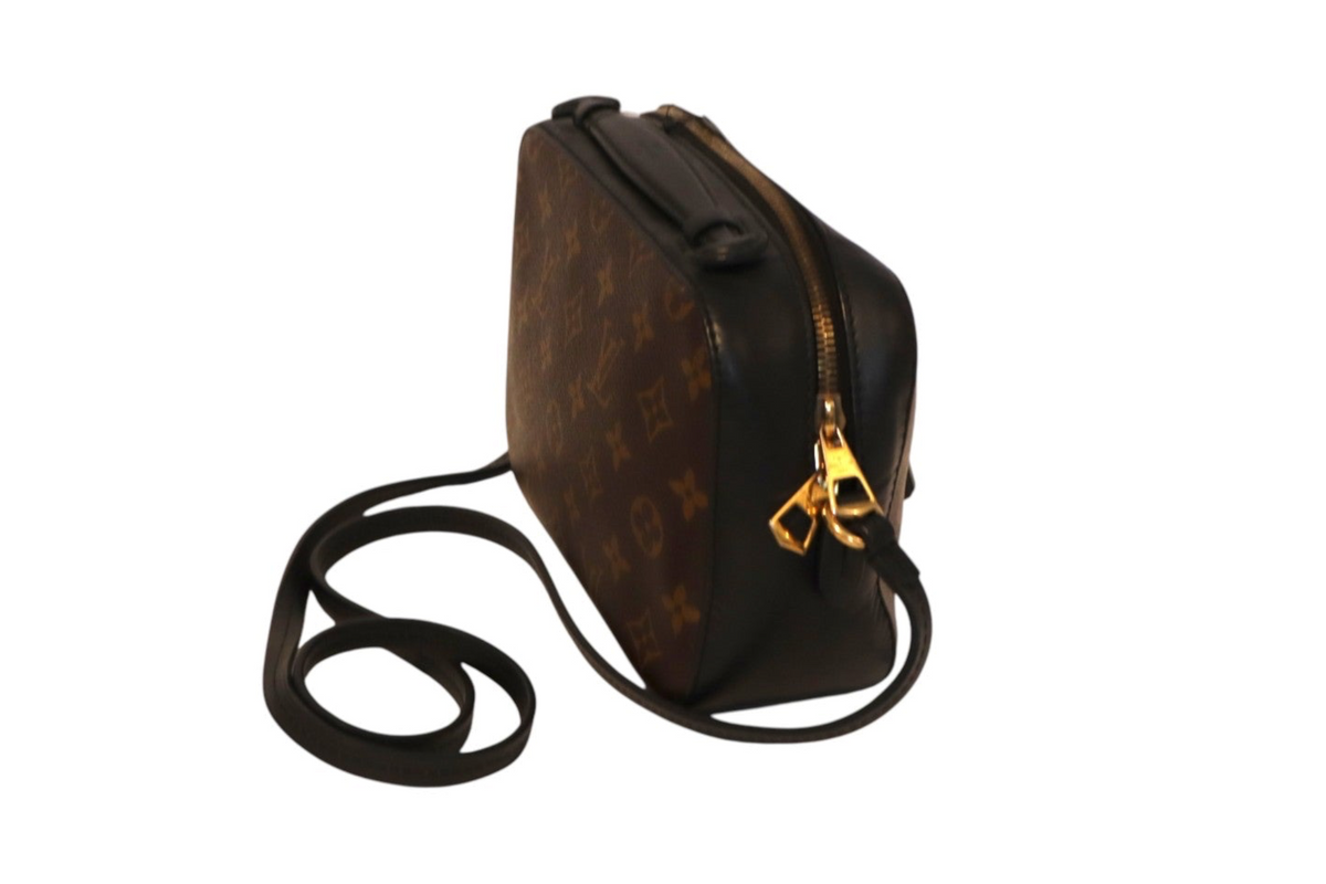 Side angle view of Louis Vuitton Monogram canvas crossbody camera bag with black leather trim, gold-tone zipper hardware, and long black leather shoulder strap on a white background.