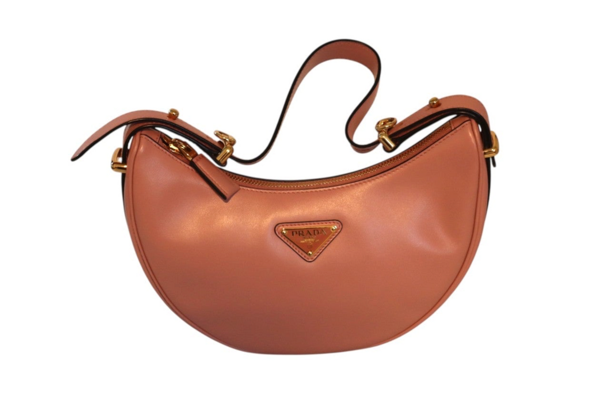 A Prada crescent-shaped leather shoulder bag in pink, featuring gold-tone hardware and the signature triangular Prada logo plaque on the front.
